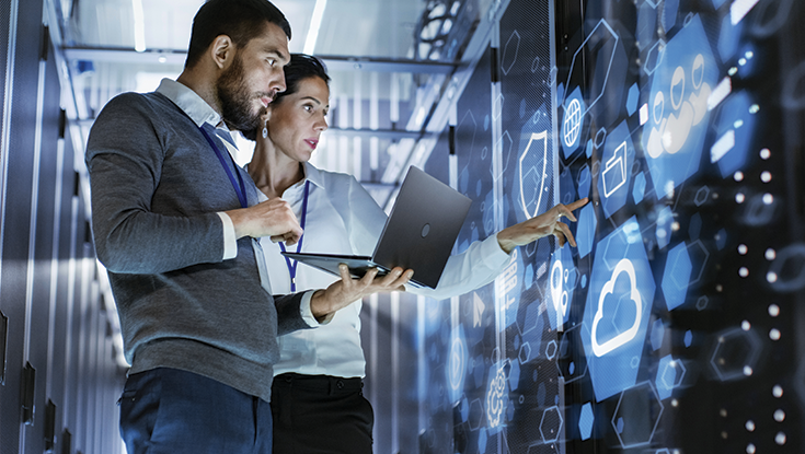 Two professionals, one holding a laptop, discussing in front of a data dashboard